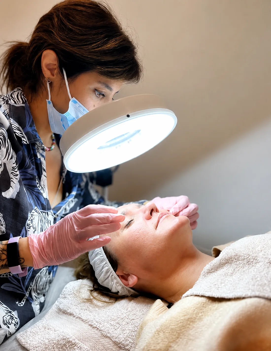 Cande, con barbijo y guantes, hace una limpieza facial a una clienta bajo una lámpara lupa