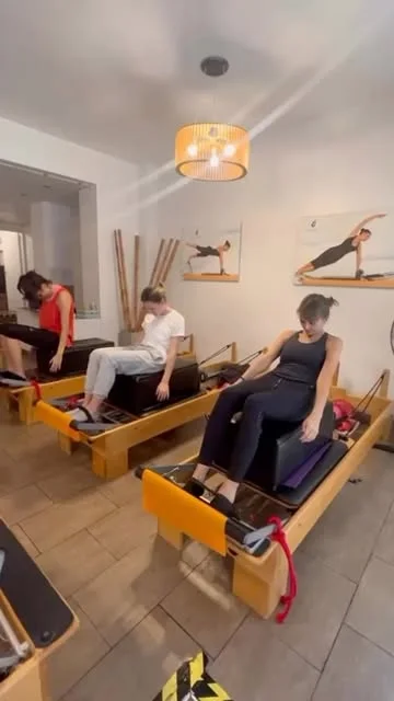 Tres alumnos en reformers durante una clase, con cuadros de posturas de pilates en la pared