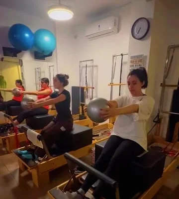 Alumna sentada en el reformer sosteniendo una pelota con los brazos extendidos