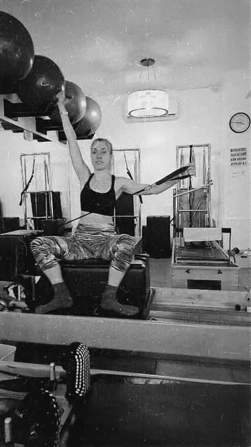 La profe Nayla sentada en un reformer con los brazos en las cintas, foto en blanco y negro