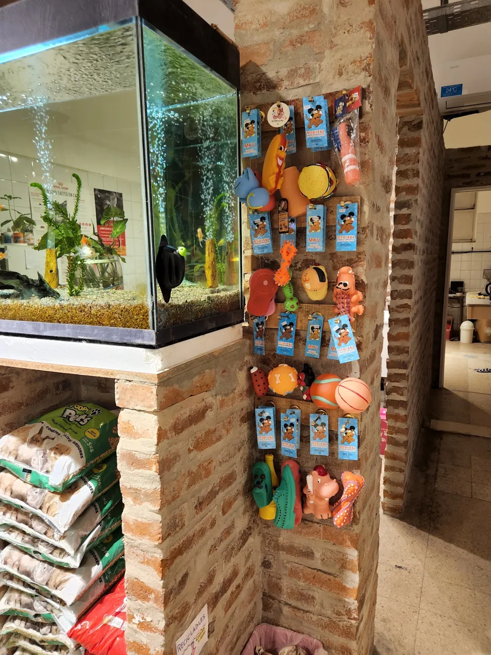 Interior de la veterinaria: pecera sobre bolsas de alimento y una columna de ladrillo con juguetes para perros colgados